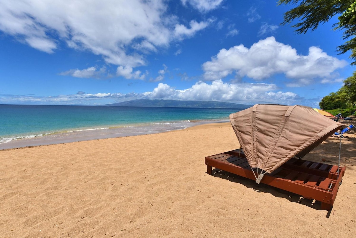A tranquil beach scene features golden sand under a vast blue sky, dotted with white clouds. A wooden lounge chair is positioned on the sand, with a large sunshade providing shelter. Gentle waves lap at the shoreline, completing the serene coastal view.