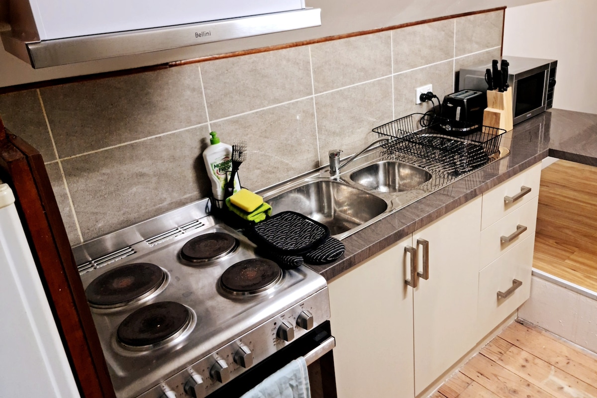 A functional kitchen area is equipped with a four-burner stove, a two-basin sink, and a countertop microwave. Counter storage holds a dish rack, a toaster, and utensils, with a bottle of dish soap placed by the sink. Wooden flooring enhances the space.