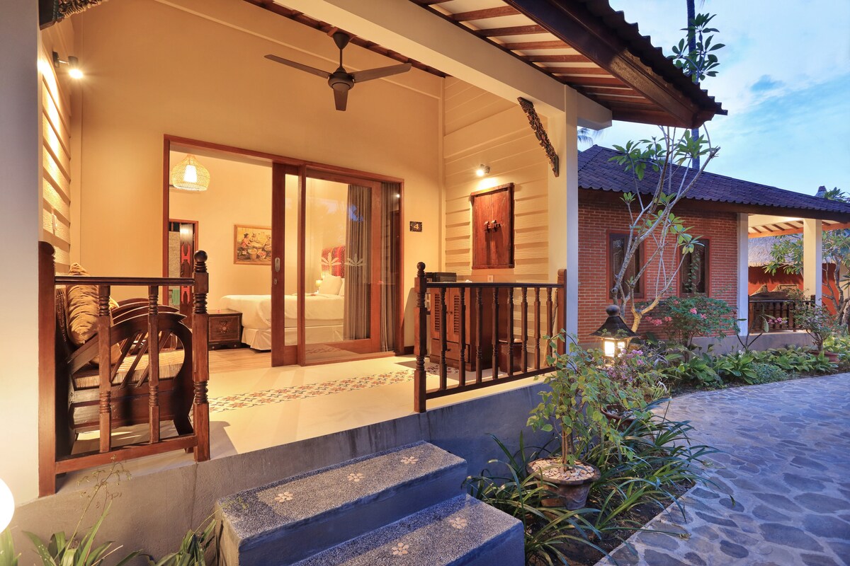 The entrance to a villa is surrounded by lush greenery and stone pathways. A spacious patio area includes wooden seating, while large sliding doors lead into a brightly lit interior. The villa's garden features tropical plants, creating a serene and inviting atmosphere.