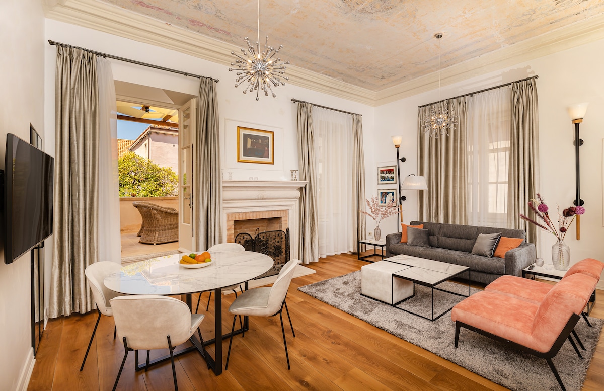 The living area features a blend of modern and classic styles, with a spacious grey sofa and an orange accent chair arranged around a light rug. A round dining table echoes the room's sophistication, while frescoed ceilings and oversized windows offer a glimpse of natural light.