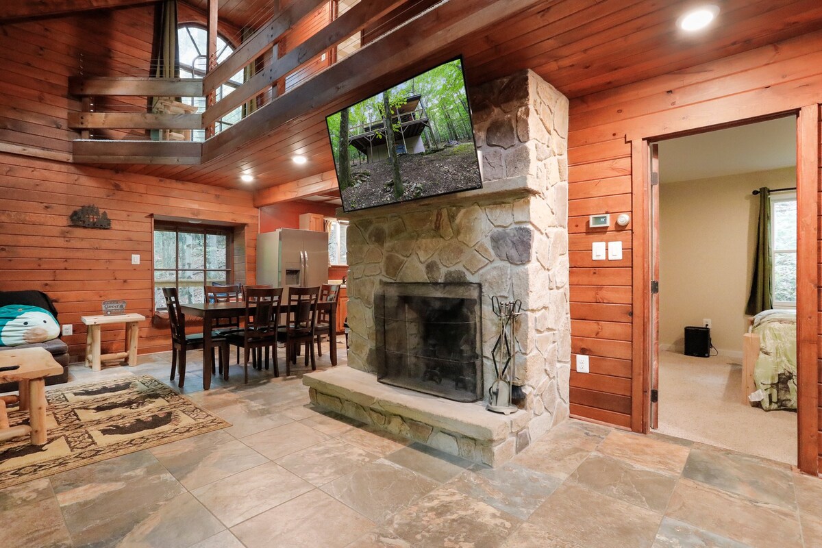 The open living space features a stone fireplace, a wooden dining table with seating, and a mounted television. Natural wood paneling and large windows provide a warm atmosphere. Slate tile flooring extends throughout, while a doorway leads to a cozy bedroom area.