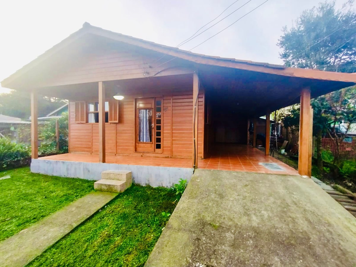 A wooden chalet features a spacious covered porch supported by wooden pillars. The entrance is accentuated by large windows and wooden shutters. A concrete pathway leads to the front door, surrounded by a well-maintained grassy area and a gentle slope leading to the side.