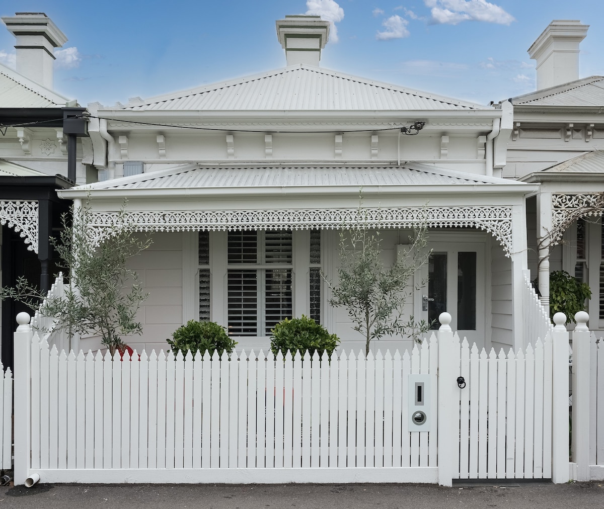 The exterior of a charming Victorian-style home is presented, featuring a white picket fence and a decorative front porch. Olive trees are positioned on either side of the entrance, adding greenery to the façade. The building showcases classic architectural details and a welcoming look.