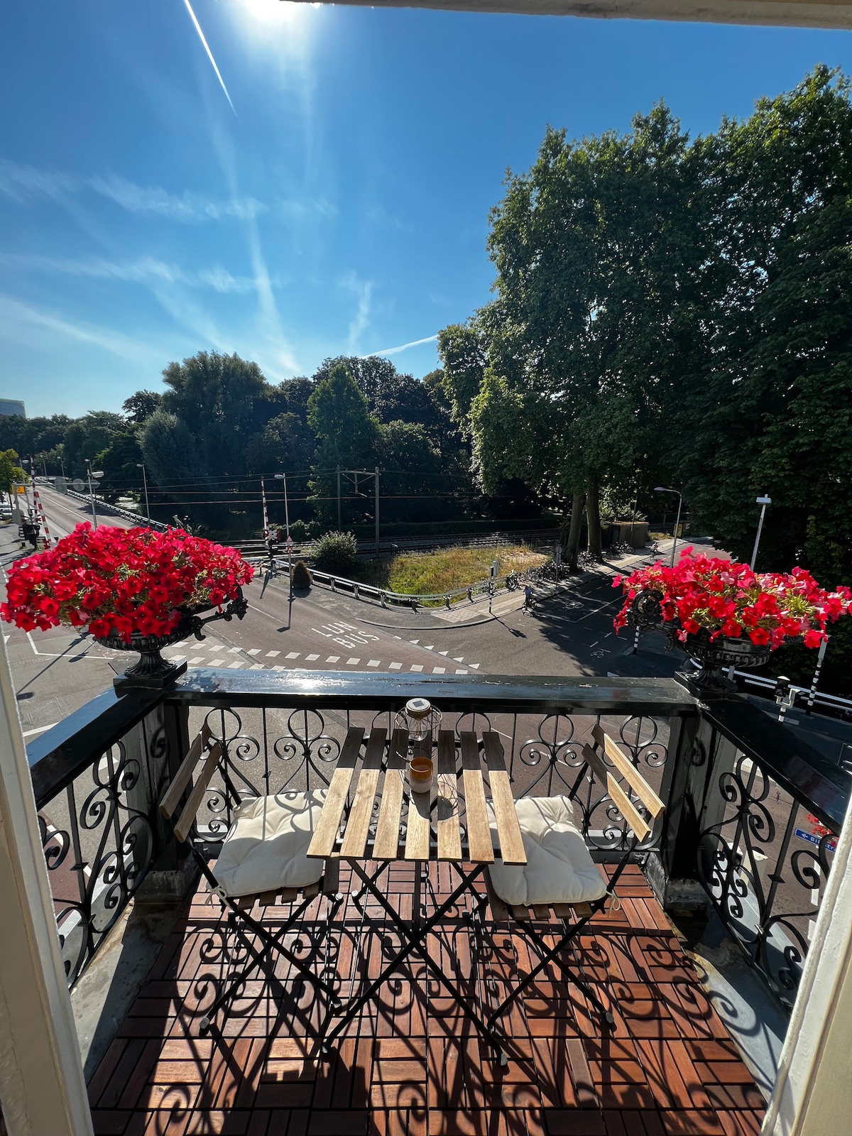 A balcony offers a view of a tree-lined street, with vibrant red flowers adorning the railing. A small wooden table and two chairs are positioned on the tiled floor, inviting relaxation. Sunlight illuminates the scene under a clear blue sky.