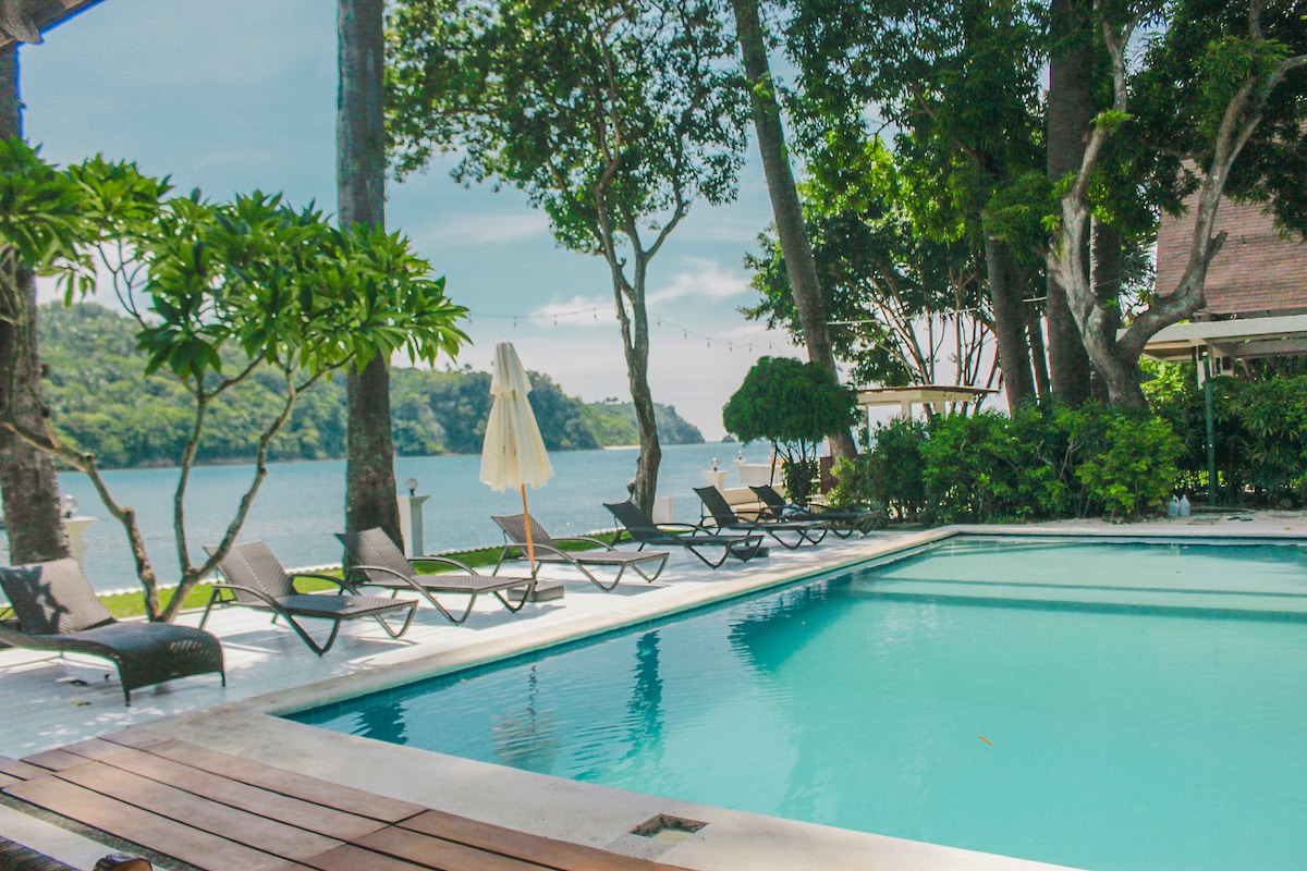 An outdoor swimming pool is bordered by lounge chairs under the shade of surrounding trees. A large umbrella provides sun protection beside the pool. The tranquil water reflects the distant landscape, enhancing the relaxed atmosphere of the garden villa setting.