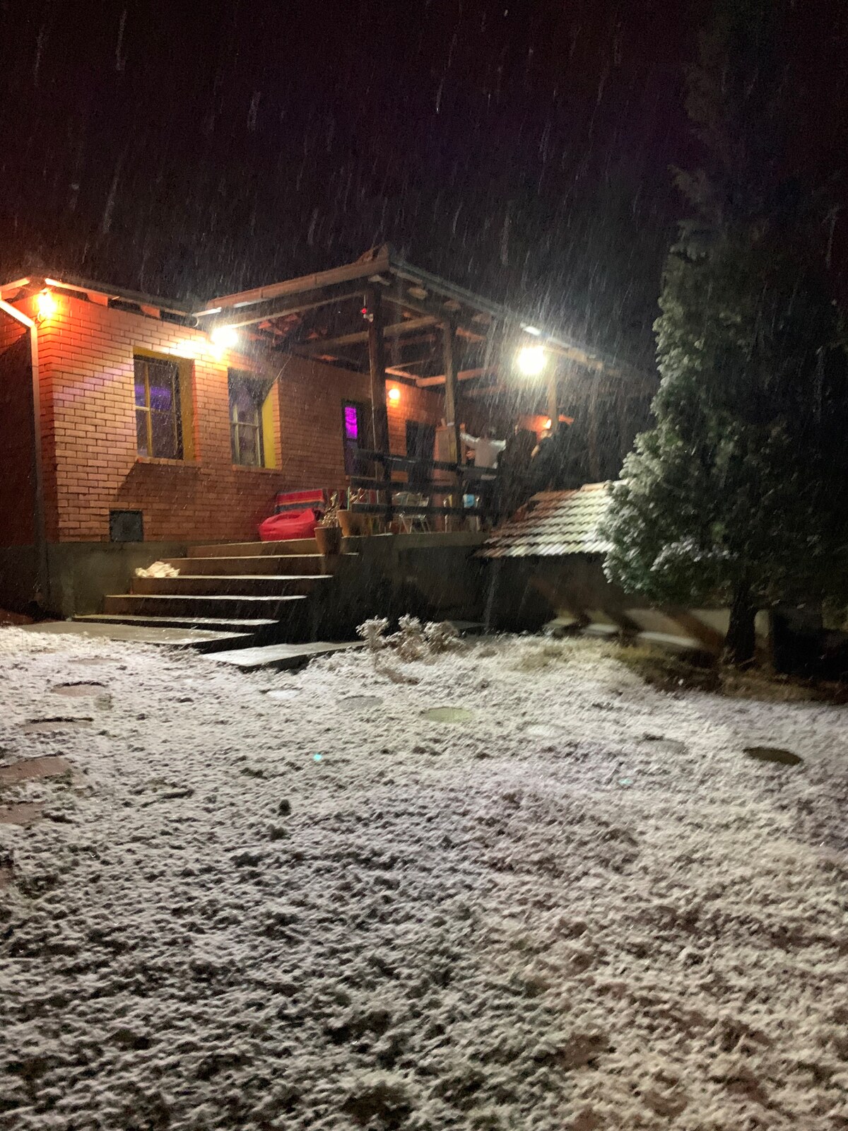 The house is illuminated by warm outdoor lights, surrounded by falling snow. A set of stairs leads up to a covered porch, furnished for relaxation. The snowy landscape adds a serene touch to the cozy cabin setting.