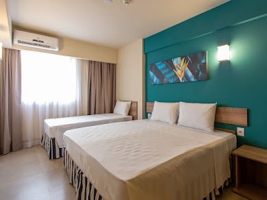 A bright guest room features a spacious bed alongside a single bed, both dressed in crisp white linens. A colorful wall art piece adds a touch of style. Natural light filters in through a large window, complemented by soft curtains.