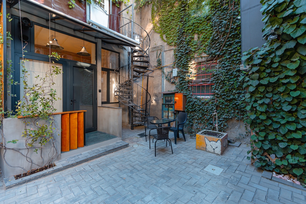 An outdoor courtyard is surrounded by lush green ivy-covered walls, featuring a spiral staircase leading to upper levels. A small table with chairs is positioned on the stone-paved surface, while a vintage-style telephone booth adds a unique touch to the space.