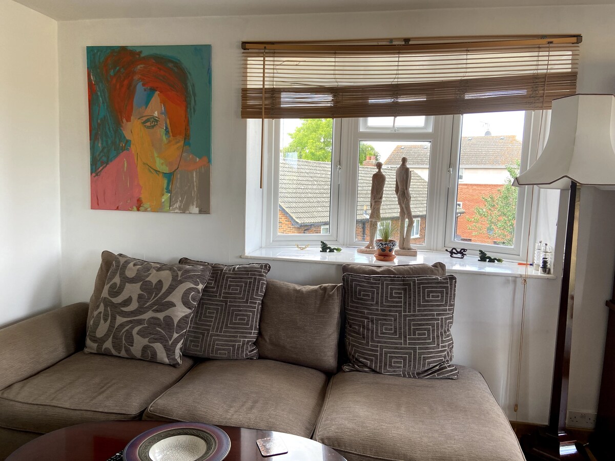 A comfortable seating area is highlighted by a sectional sofa adorned with patterned cushions. Natural light filters through large windows, complementing a vibrant artwork displayed on the wall. Decorative figures and plants are positioned along the windowsill, creating an inviting ambiance.