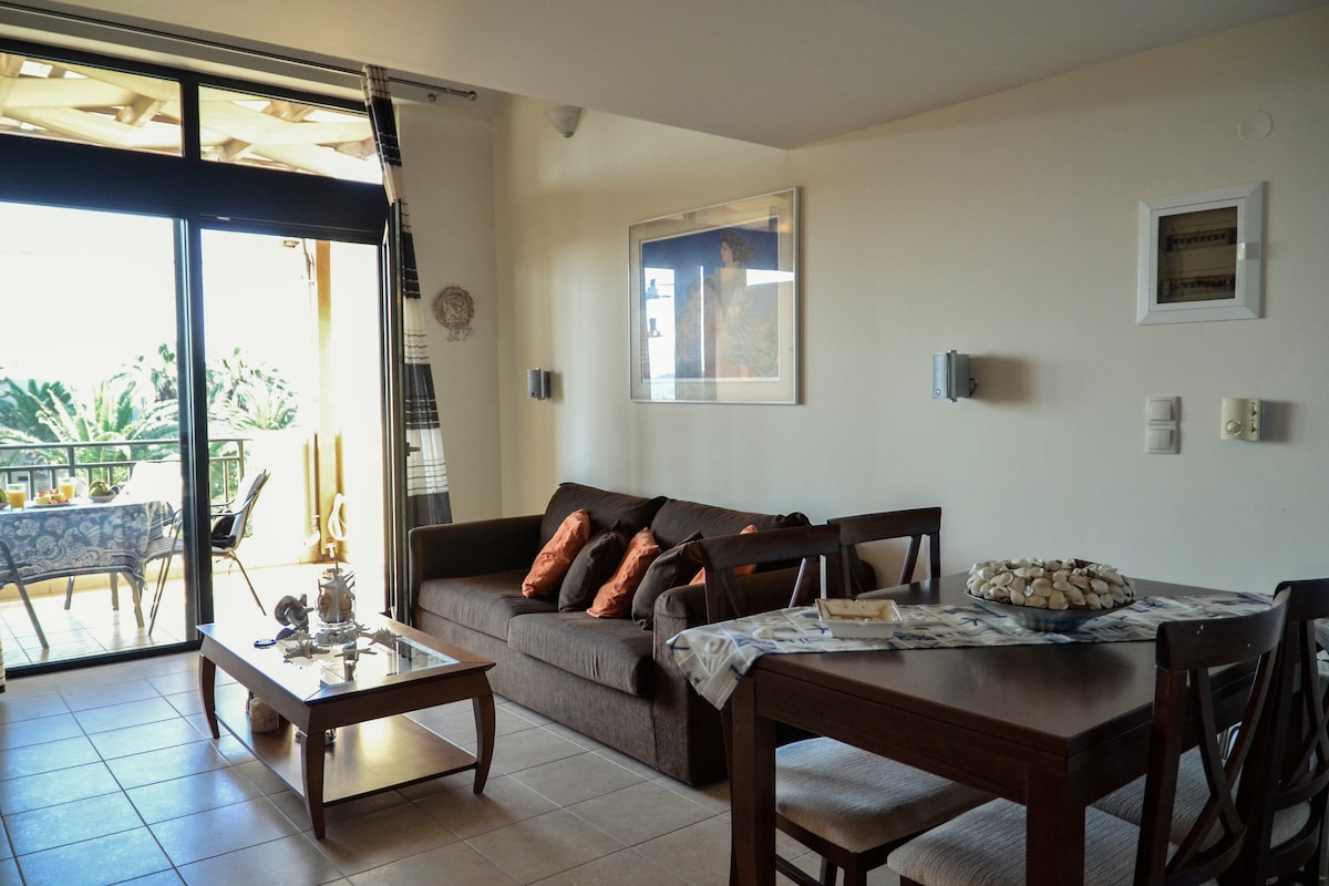 An open-plan living area features a comfortable couch with decorative pillows, a wooden dining table, and a side table displaying decorative objects. Sliding glass doors lead to a balcony, allowing natural light to fill the space and offering views of the surrounding area.