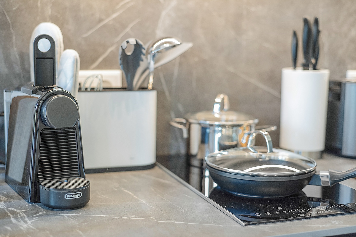 A modern kitchen countertop features a coffee machine beside a pot and frying pan. Utensils are neatly organized in a container, with additional kitchen tools visible in the background. The surface is sleek, complemented by stylish cabinetry and a minimalist design.