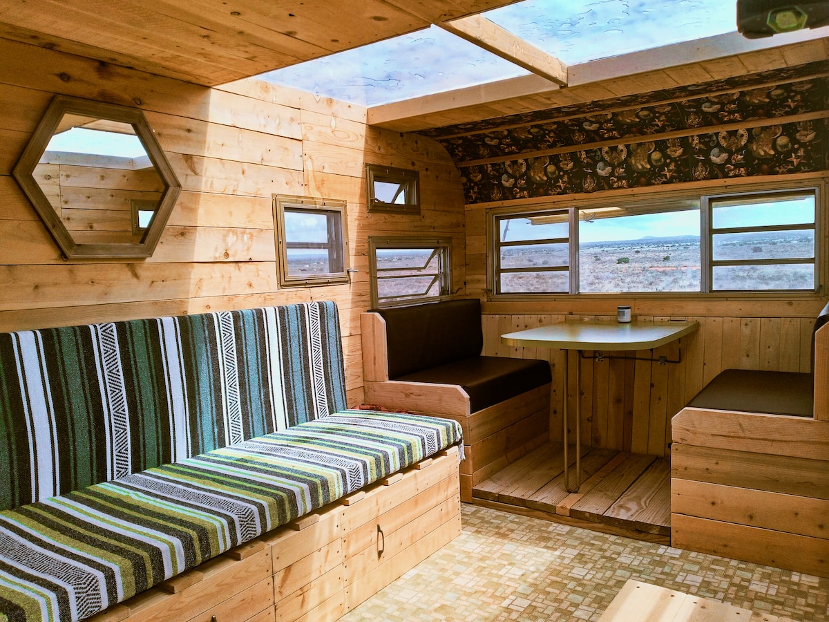 The interior of the vintage camper features wooden walls and a large skylight, allowing natural light to fill the space. Seating areas are lined with striped fabric, and a small table is set between additional seating, creating a cozy and functional layout.