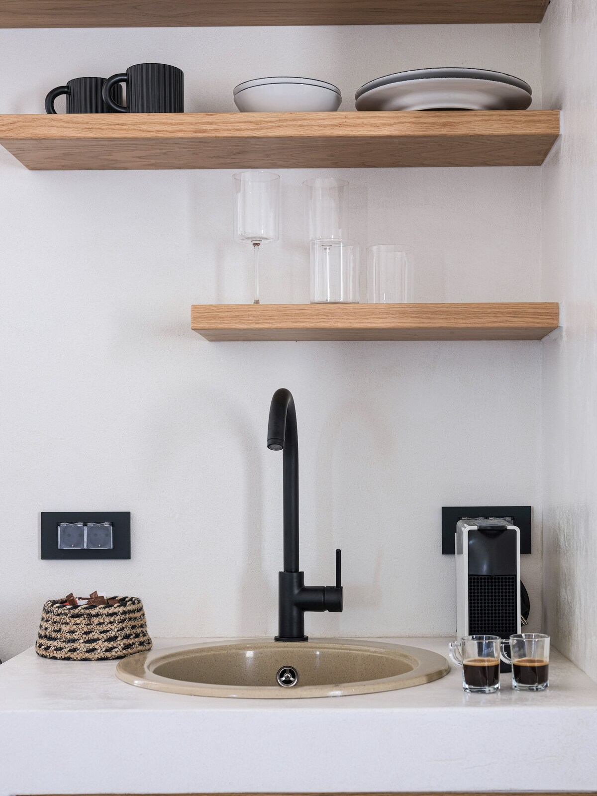 A modern kitchenette is displayed, featuring a sleek black faucet above a circular sink. Open wooden shelves hold a mix of dishware and glassware, while a coffee machine with accompanying cups adds functional elegance to the space. The countertops are minimalistic with a neutral tone.