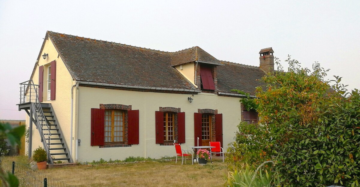 A charming cottage is showcased with a sloped roof and red shutters. An external staircase leads to a second-floor suite. The grassy area in front includes a small table and chairs, inviting outdoor relaxation surrounded by greenery.