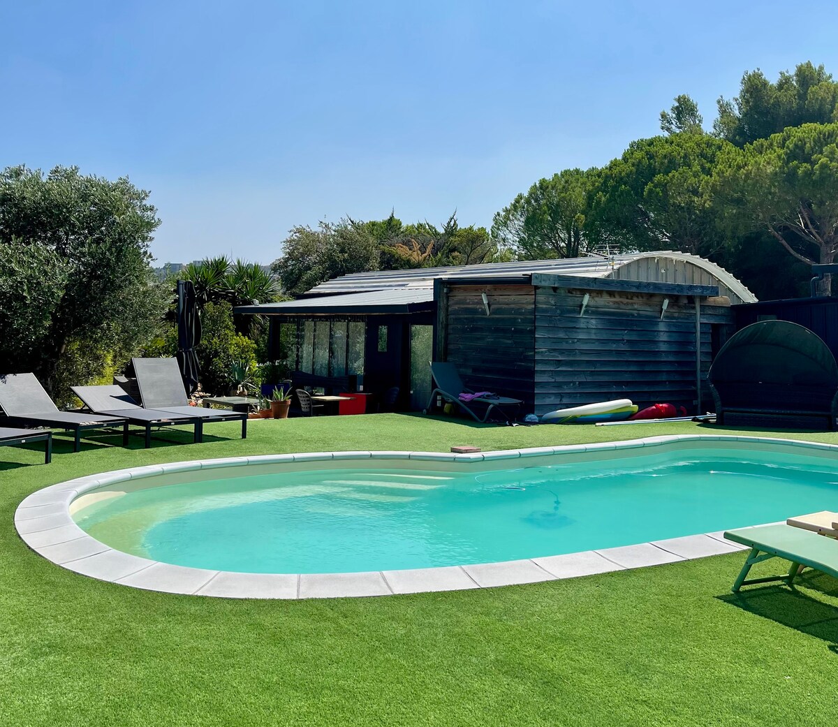 A serene outdoor space features a swimming pool surrounded by well-maintained grass. Sun loungers are positioned nearby, enhancing the relaxation area. A wooden structure with expansive windows sits in the background, seamlessly integrated into the tranquil environment.
