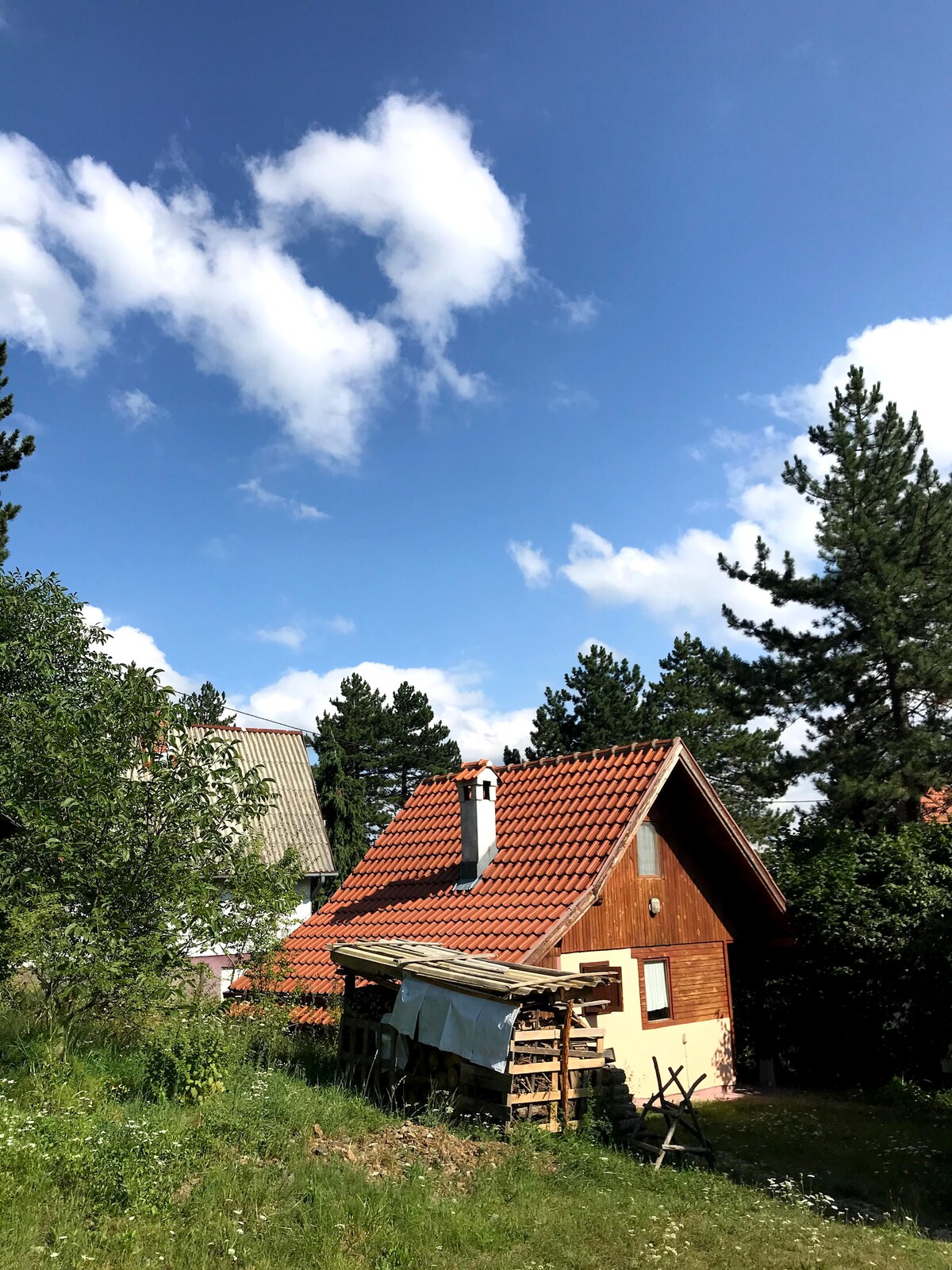A wooden bungalow with a red tiled roof is nestled among lush greenery under a clear blue sky. The structure features a chimney and is partially surrounded by trees, conveying a serene natural setting.