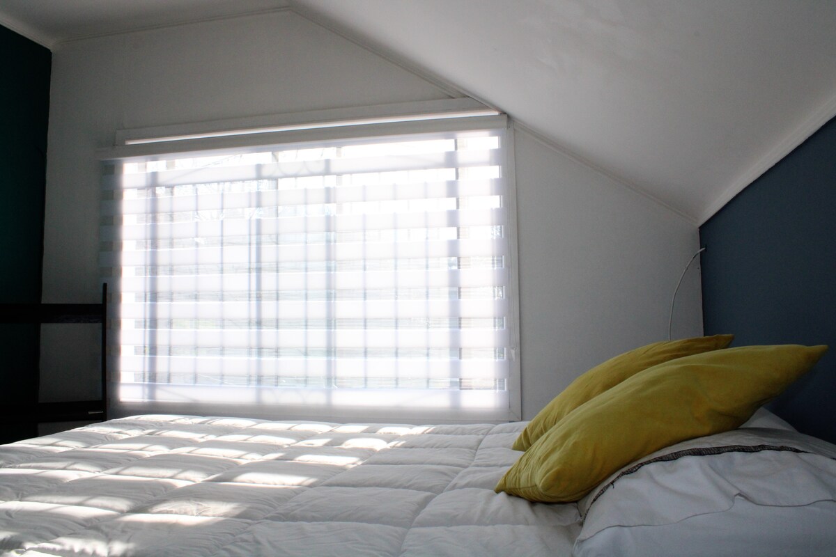 A sunlit bedroom features a spacious bed with white bedding and two yellow accent pillows. Natural light filters through a large window adorned with sheer blinds, casting gentle shadows across the bed. The walls are painted in calming shades of blue and white.