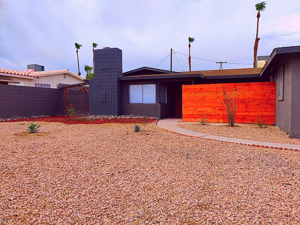 The exterior of the home showcases a combination of modern design with a grey façade and a prominent orange wooden wall. The landscaped yard features gravel with scattered low-maintenance plants and a pathway leading to the entrance.