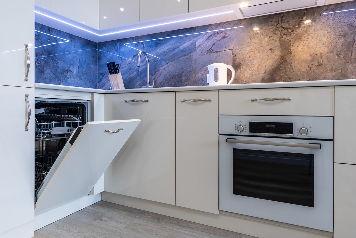 A modern kitchen is showcased with sleek cabinetry and a stone backsplash. A built-in dishwasher with an open door reveals its interior, while an oven is integrated below a countertop. A kettle and utensils are positioned neatly on the counter, illuminated by soft lighting.