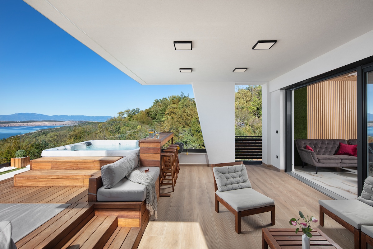 An outdoor terrace features a hot tub and a seating area, surrounded by wooden lounge furniture. Large sliding glass doors open to a living room, and a panoramic view of the landscape with trees and water stretches in the background.