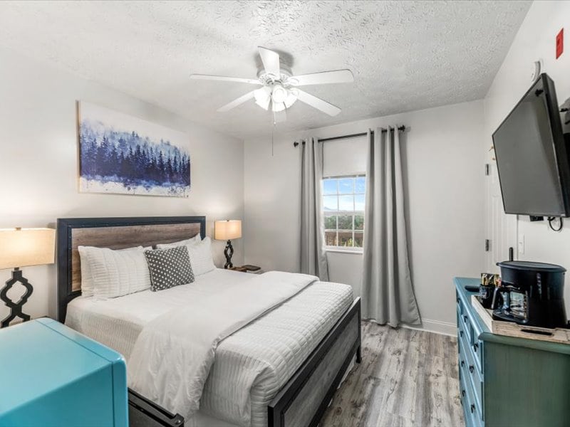 A well-lit bedroom features a spacious bed with soft linens and decorative pillows. A fan suspended from the ceiling provides airflow. Beside the bed are two lamps on nightstands, and a television is mounted on the opposite wall. Natural light enters through a window covered by light curtains.