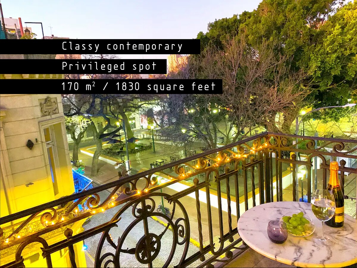 A small balcony is viewed at dusk, featuring a round marble table with a glass of wine and fresh fruit. Decorative lights illuminate the space, offering a glimpse of the vibrant plaza below, surrounded by trees with bare branches.