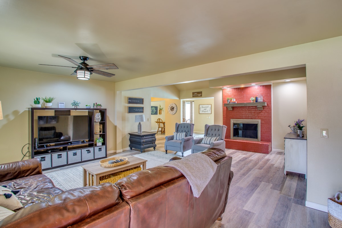 The living room features a comfortable sectional sofa, two accent chairs, and a coffee table. A wall-mounted TV is positioned across from the seating area, and a fireplace is set within a brick wall. Natural light filters in, enhancing the inviting space.