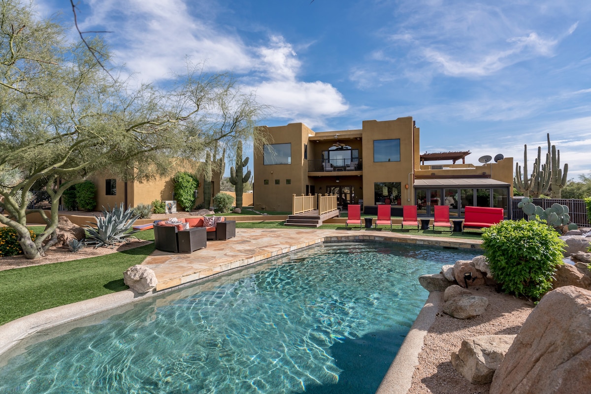 A private backyard is depicted, featuring a heated pool surrounded by a paved patio. Lounge chairs provide seating options under clear skies. The estate is framed by desert vegetation, with a two-story structure visible in the background, showcasing large windows and outdoor decks.