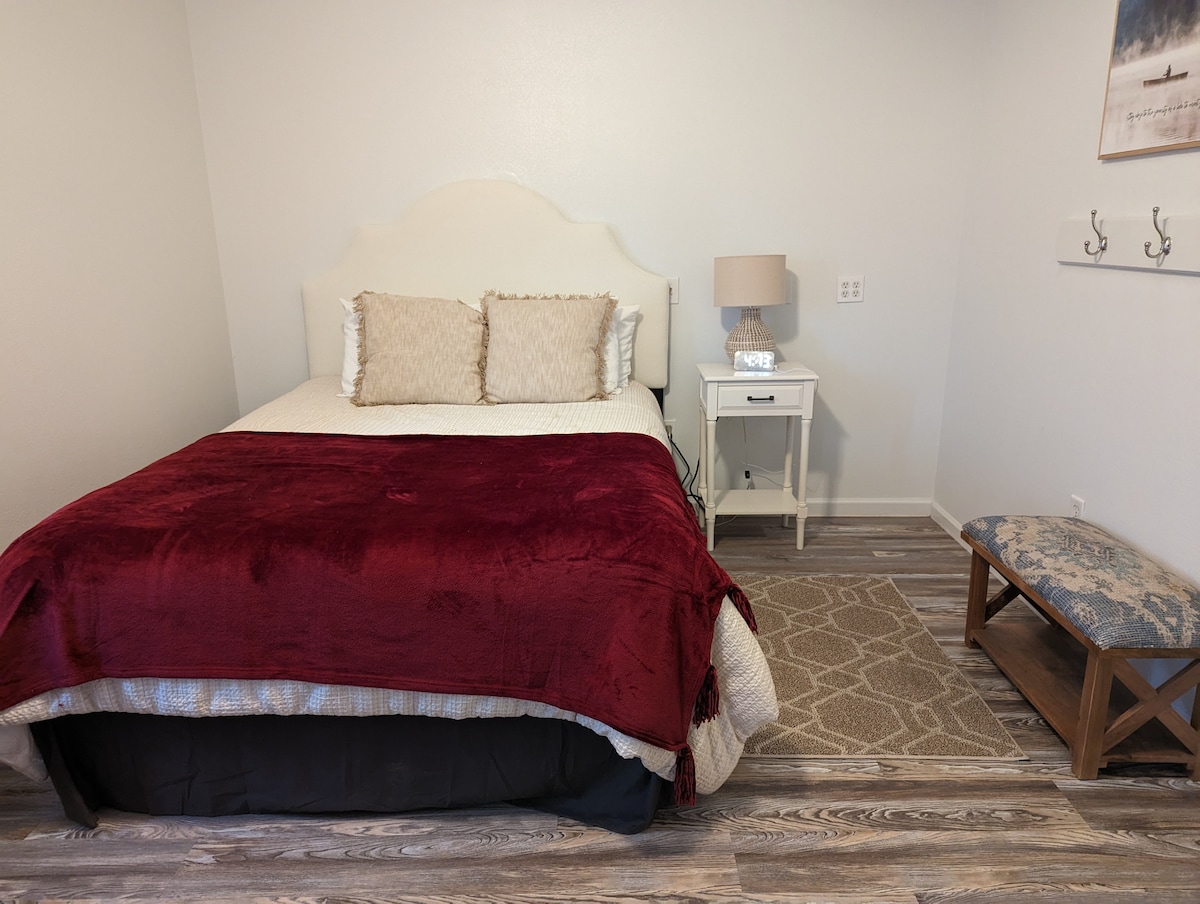 A cozy sleeping area features a spacious bed dressed in white linens with a deep red blanket draped across the foot. Two decorative pillows enhance the bedding. A bedside table with a lamp and a small bench provide additional functionality in the room.