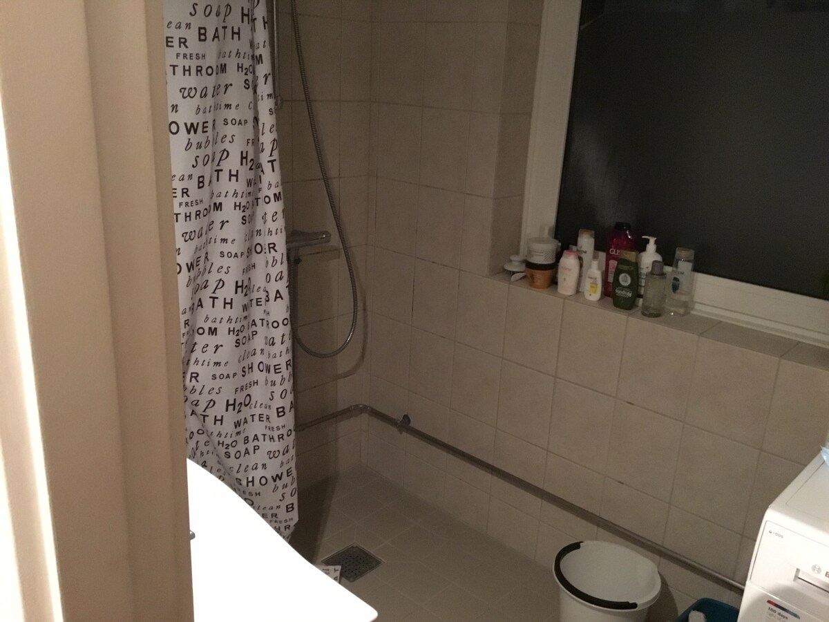 A well-equipped bathroom is shown with beige tiled walls and floor. A shower area is present, featuring a curtain with text prints. Various toiletries are arranged on a windowsill, with a washing machine visible on the side.