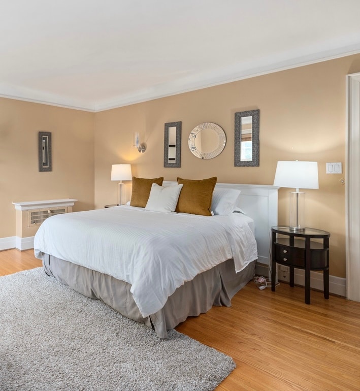 A spacious bedroom features a light-colored bed adorned with neatly arranged pillows. Two bedside lamps provide soft lighting on either side of the bed. A plush area rug covers the wooden floor, and a series of mirrors are mounted on the wall, enhancing the room's brightness.