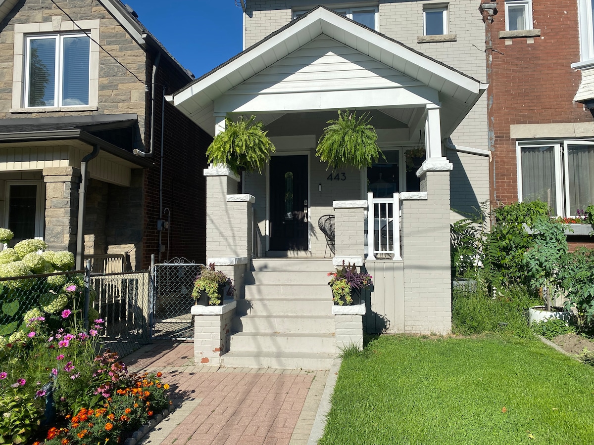 The entryway features a covered porch adorned with two hanging ferns, framed by light-colored brick. A welcoming front door is set between white columns, while a path of interlocking bricks leads to the stairs, which are flanked by planters filled with vibrant flowers.