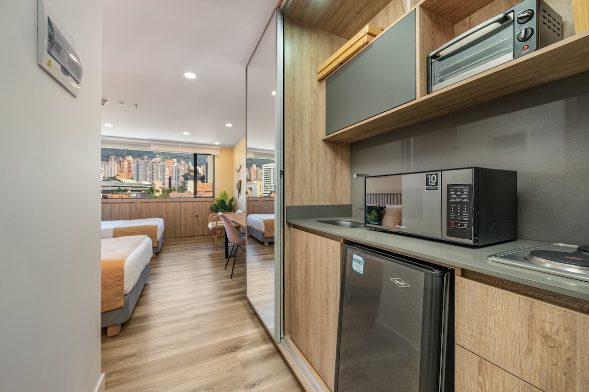 A kitchenette is visible, featuring a small fridge, microwave, and cooktop, with wooden cabinetry above. The spacious living area, separated by sliding mirror doors, showcases two double beds and large windows, providing a view of the city beyond.
