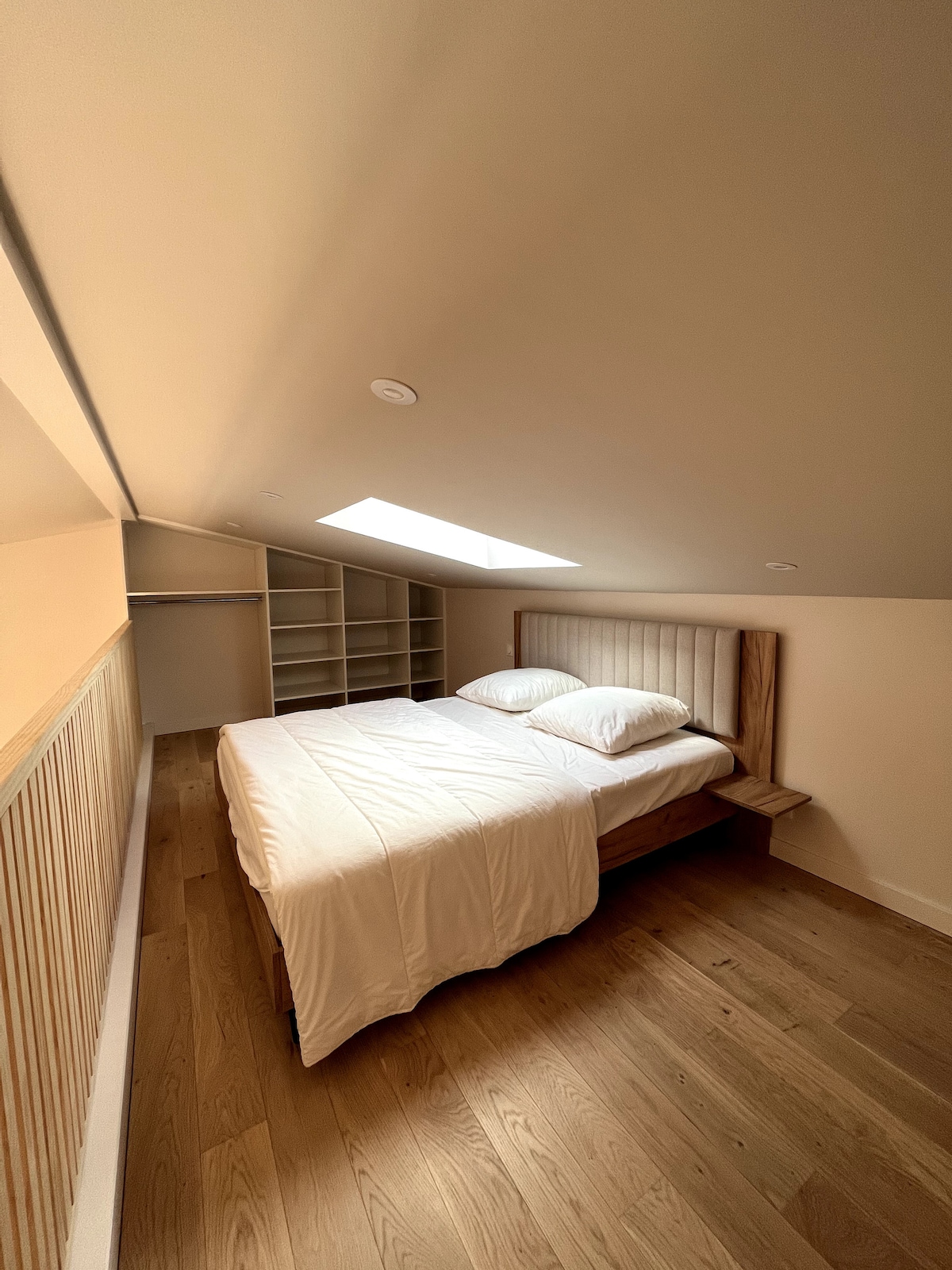 A cozy sleeping area is featured, with a spacious bed dressed in white linens. Natural light enters through a skylight, illuminating the wood floor. Shelving units are visible in the background, providing additional storage options.