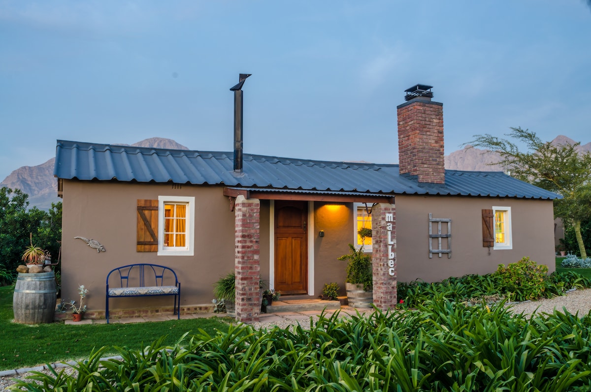The cottage features a charming exterior with a rustic brick chimney and a dark metal roof. Two windows are framed with wooden shutters, while a welcoming wooden door leads inside. A small bench sits outside, surrounded by lush greenery and mountain views in the background.
