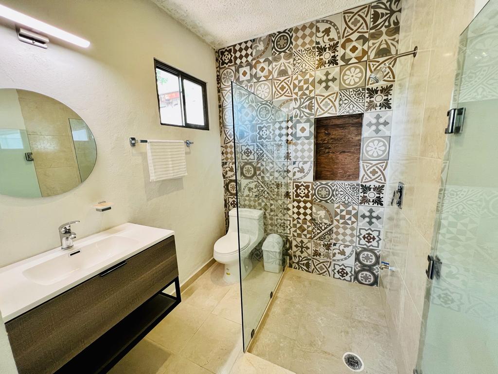 A modern bathroom features patterned tile accents on one wall. A glass shower enclosure is present, along with a stylish vanity featuring a round mirror. Natural light enters through a small window, illuminating the sleek fixtures and neutral color palette.