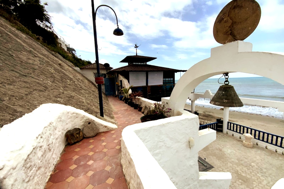 A coastal pathway is presented, featuring a smooth stone surface leading towards a building topped with a distinctive roof. Lush plants are arranged in pots along the way, and the ocean can be seen in the background, enhancing the serene atmosphere of the location.