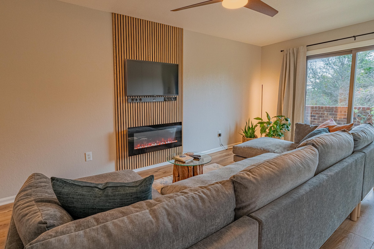 The living area features a spacious sectional sofa with soft cushions, positioned opposite an electric fireplace. A wall-mounted television is above the fireplace, surrounded by warm wood paneling. Natural light filters in through sliding glass doors, which lead to a patio surrounded by greenery.