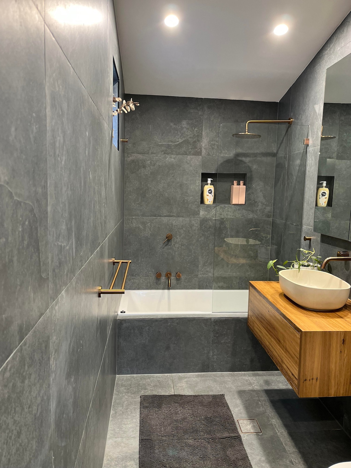 A contemporary bathroom features a soaking tub alongside a wood vanity with a bowl sink. The walls are lined with sleek dark tiles, and stylish fixtures in gold tone add an elegant touch. Soft lighting illuminates the space, enhancing the modern design.