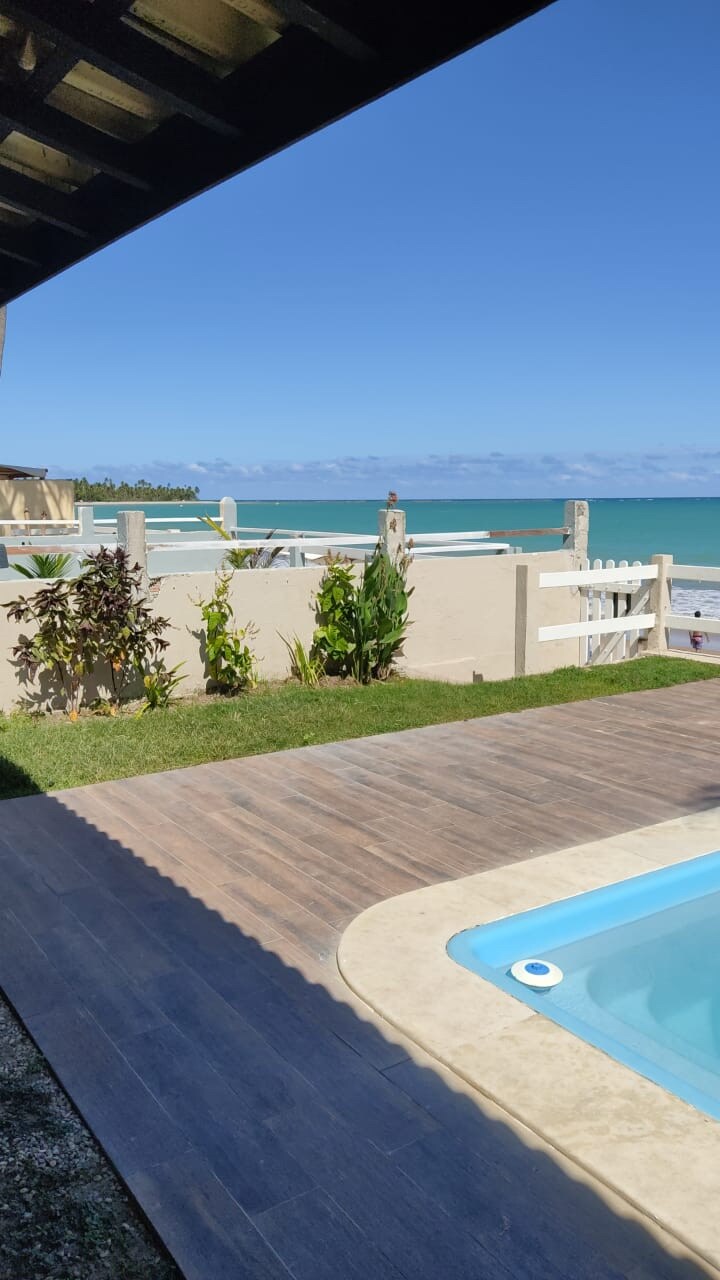 A view of the beach is visible from the garden area, showcasing lush greenery and well-maintained grass. The clear blue ocean stretches into the horizon, complemented by a bright sky. A small pool area can be seen, providing a relaxing outdoor space.