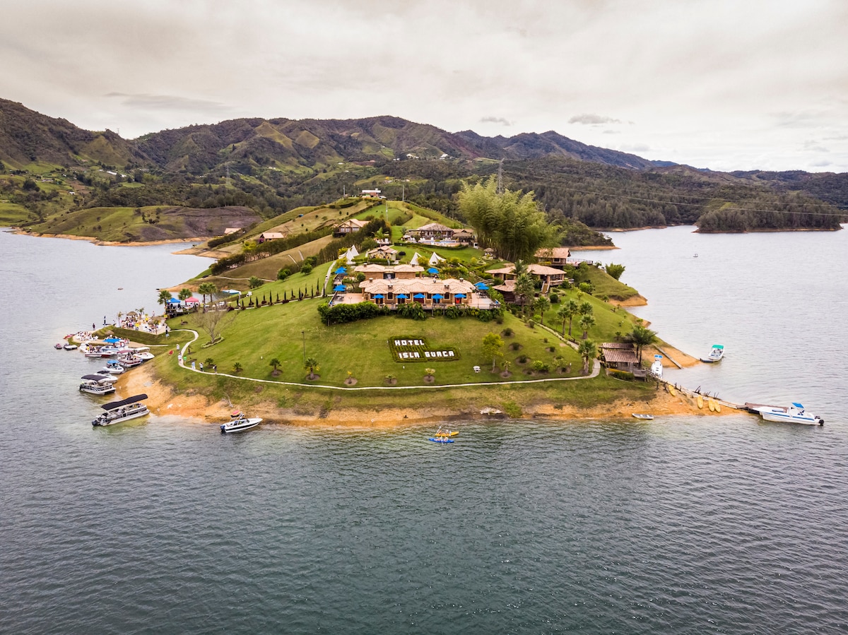 An aerial view of the island showcases a green landscape bordered by water, with a cluster of buildings and vibrant umbrellas. The natural surroundings include hills in the background, complementing the serene lakefront setting.