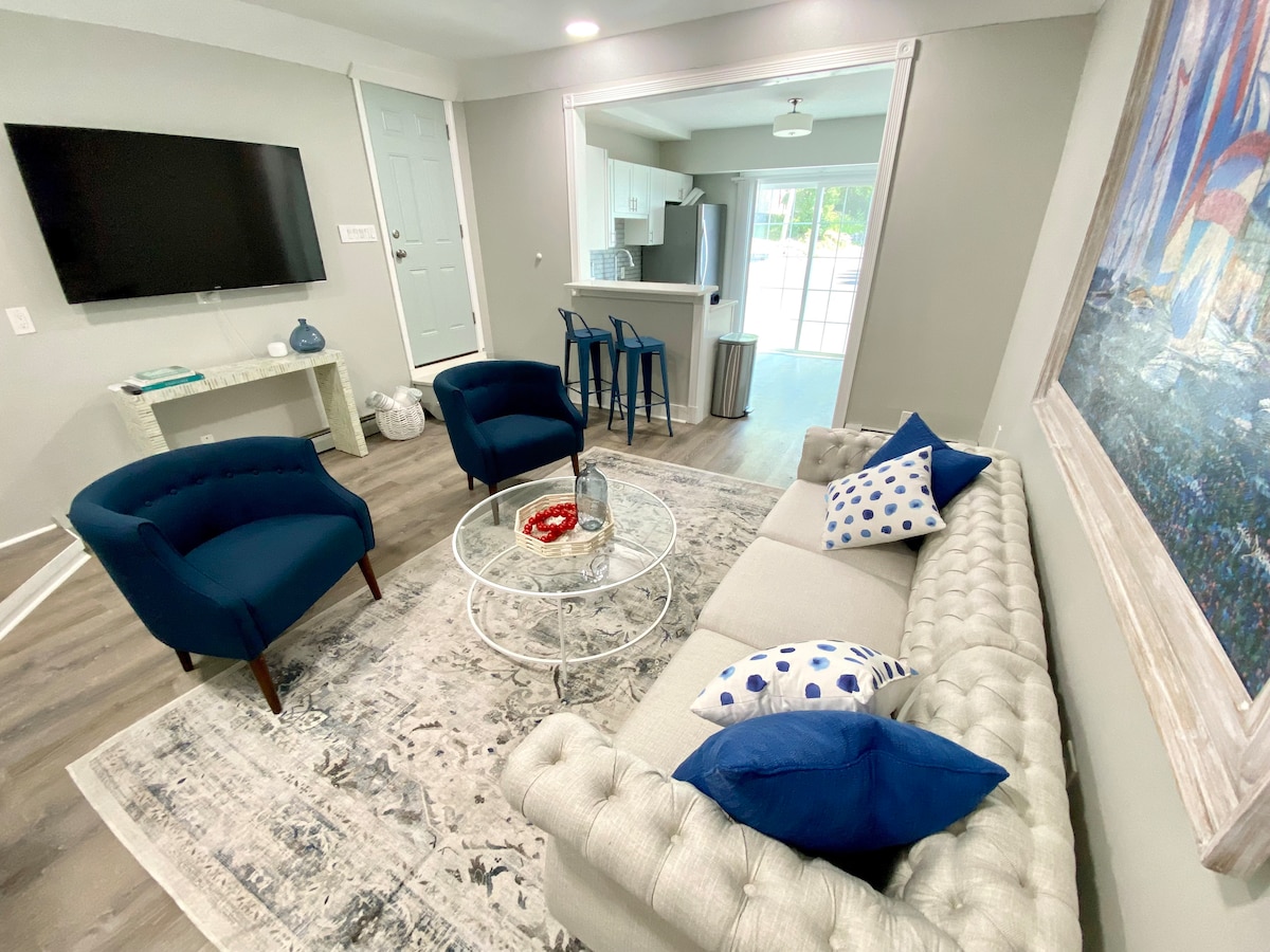 A comfortable living area features a tufted sofa adorned with blue and white cushions. A glass coffee table is centered beneath a large, colorful piece of wall art. Two blue accent chairs and a TV on the wall contribute to an inviting ambiance.