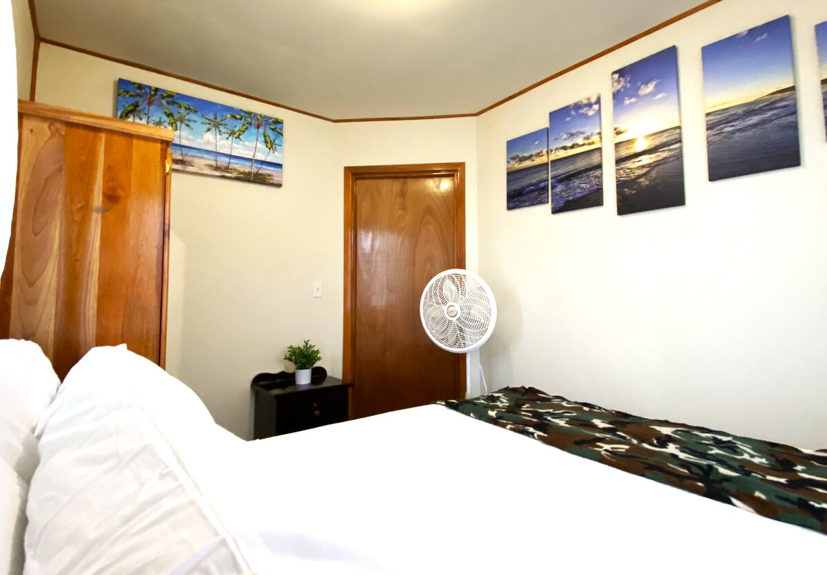 A cozy sleeping area features a spacious bed with white linens and a camouflaged throw. A small black nightstand and a decorative plant are present next to the bed. The wall is adorned with a series of framed beach landscape photos, and a standing fan is positioned nearby.