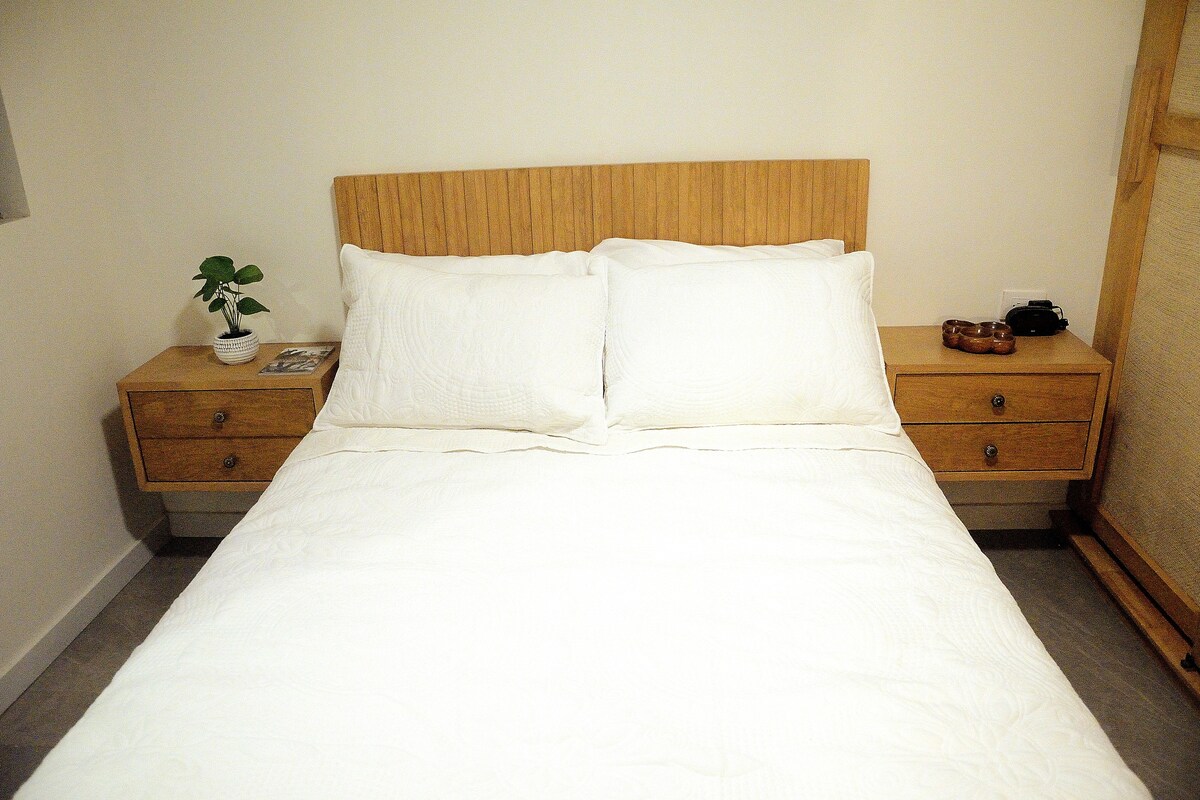A spacious bed is centered against a light wood headboard, flanked by two bedside tables. Crisp white bedding creates a clean look, and a small potted plant is present on one table. Natural light may be filtering in from an unseen window.