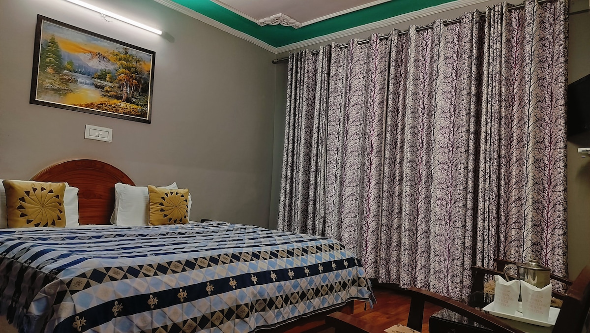 A spacious guest room features a large bed with a blue and white patterned blanket. Two white pillows are placed against a wooden headboard. Elegant curtains with a floral design frame the window, complemented by a painting of a serene landscape on the wall.