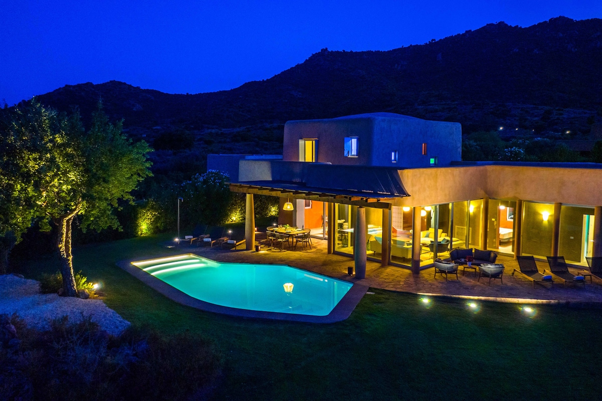 The villa is illuminated at night, showcasing a private pool with soft lighting. Surrounding greenery is visible, enhancing the tranquil ambiance. Large glass doors provide a view into the well-lit interior, while mountains silhouette the background under a deep blue sky.