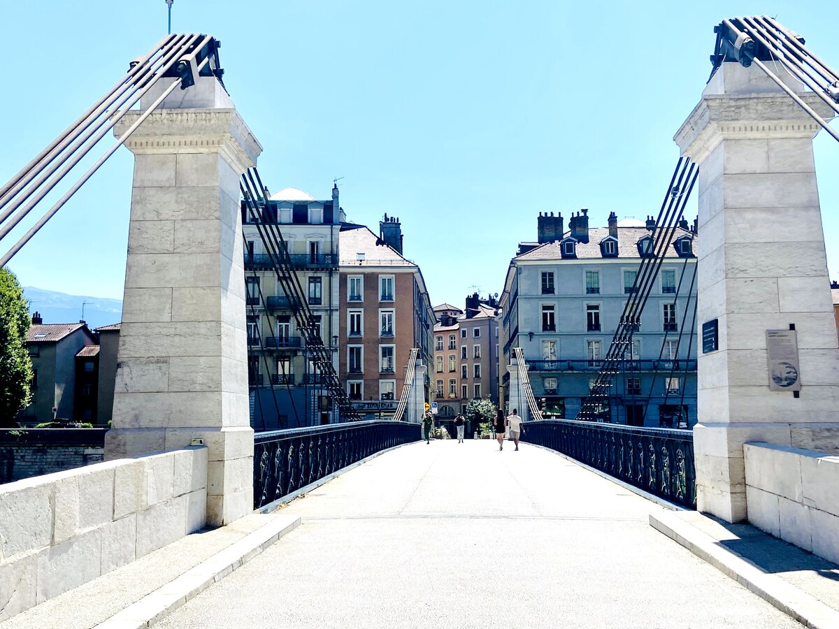 A pedestrian bridge made of stone features strong cables supporting its structure. Buildings in soft-colored facades are visible on either side, with a clear blue sky overhead. The scene captures a calm ambiance with people walking across the bridge.