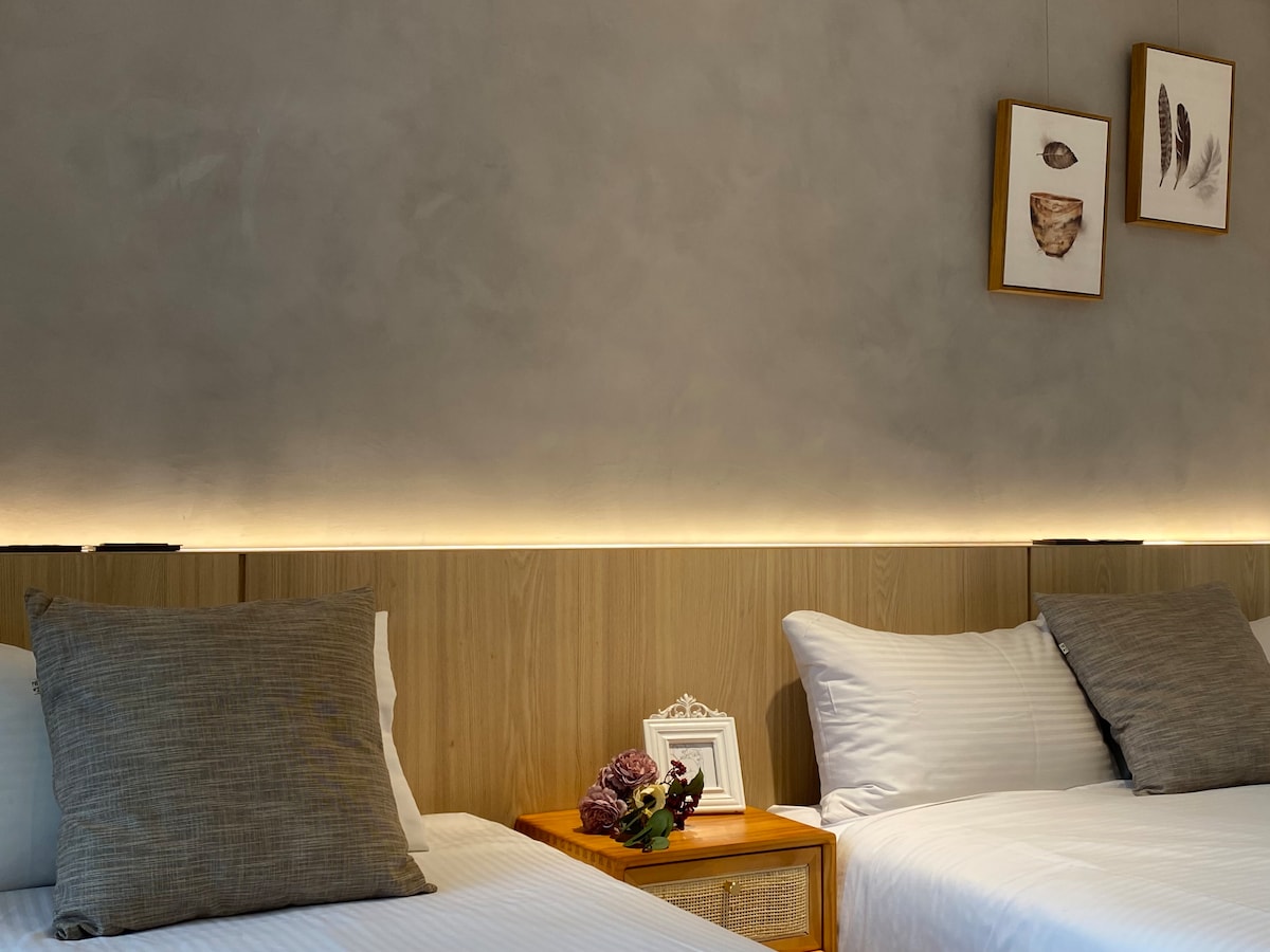 A tranquil sleeping area features two beds with light linens, complemented by soft grey decorative pillows. A wooden nightstand holds a small framed picture and some decorative flowers. The wall is adorned with simple artwork, while soft lighting enhances the peaceful ambiance.
