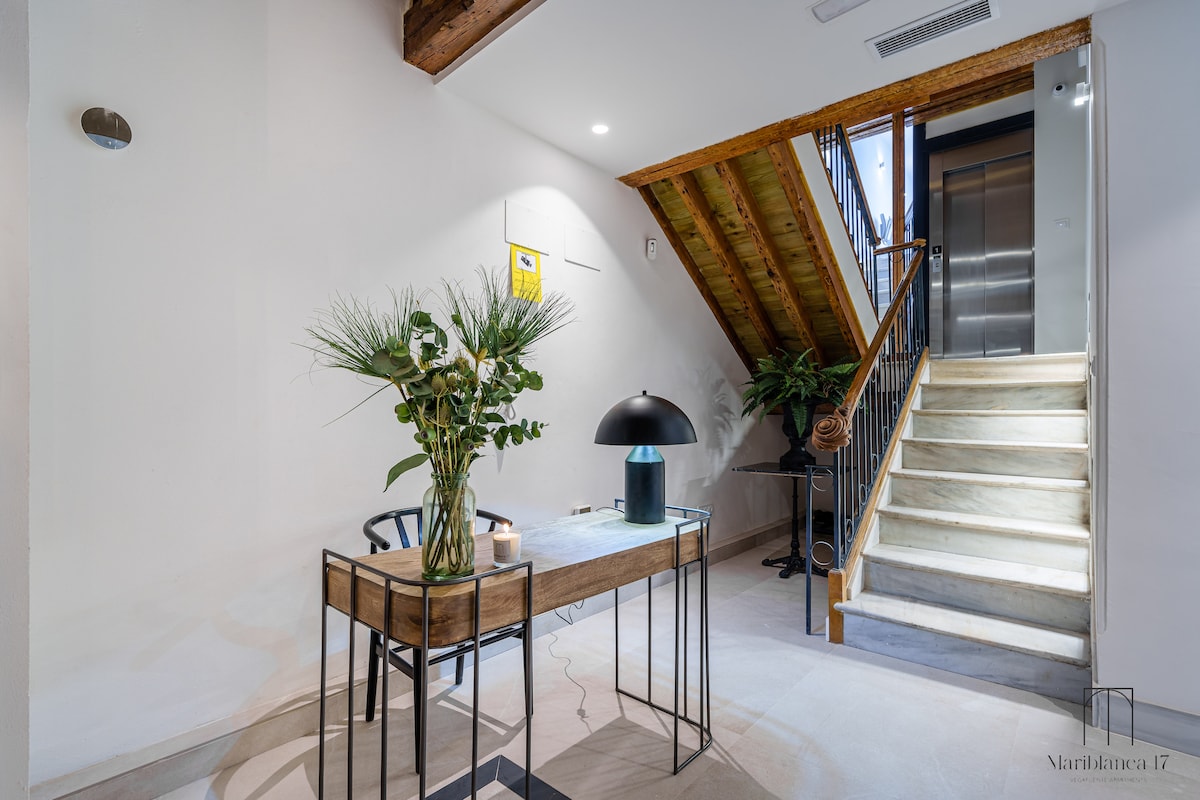 A welcoming entrance area features a wooden staircase leading to the upper floors and an elevator. A modern table is adorned with a decorative plant and a stylish lamp, enhancing the bright and inviting atmosphere.