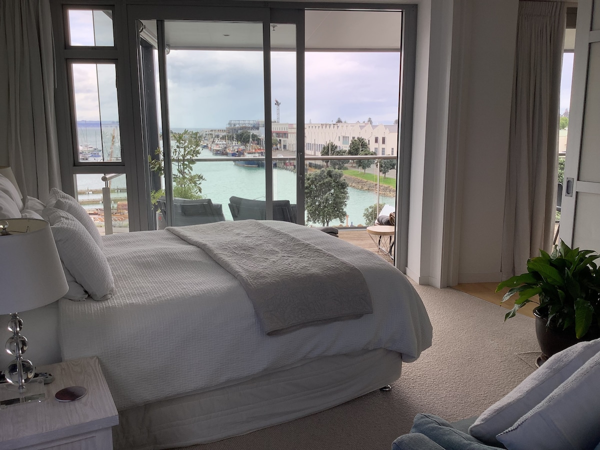 A spacious bedroom is presented with a soft color palette, offering a serene ambiance. Large glass sliding doors open to reveal stunning views of the harbor and boats. Natural light filters through, highlighting a comfortable bed and cozy seating area.